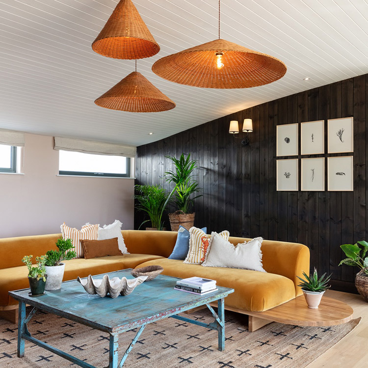 Ein stilvolles Ecksofa in einem Zimmer mit Hartboden und Teppich. Moderne Lampenschirme, ein Tisch und zahlreiche Pflanzen sind zu sehen.