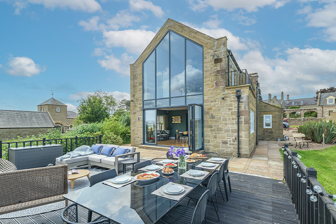 Exterior of a stone-built property with floor-to-ceiling windows. In the foreground, there's smart outdoor furniture.