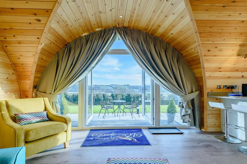 Looking out of the patio doors of a wooden glamping lodge, with decking, patio furniture and surrounding countryside.