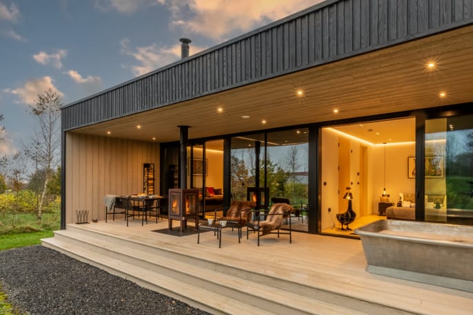 Th exterior of a large, modern lodge with floor-to-ceiling windows, a wood burner and outdoor furniture