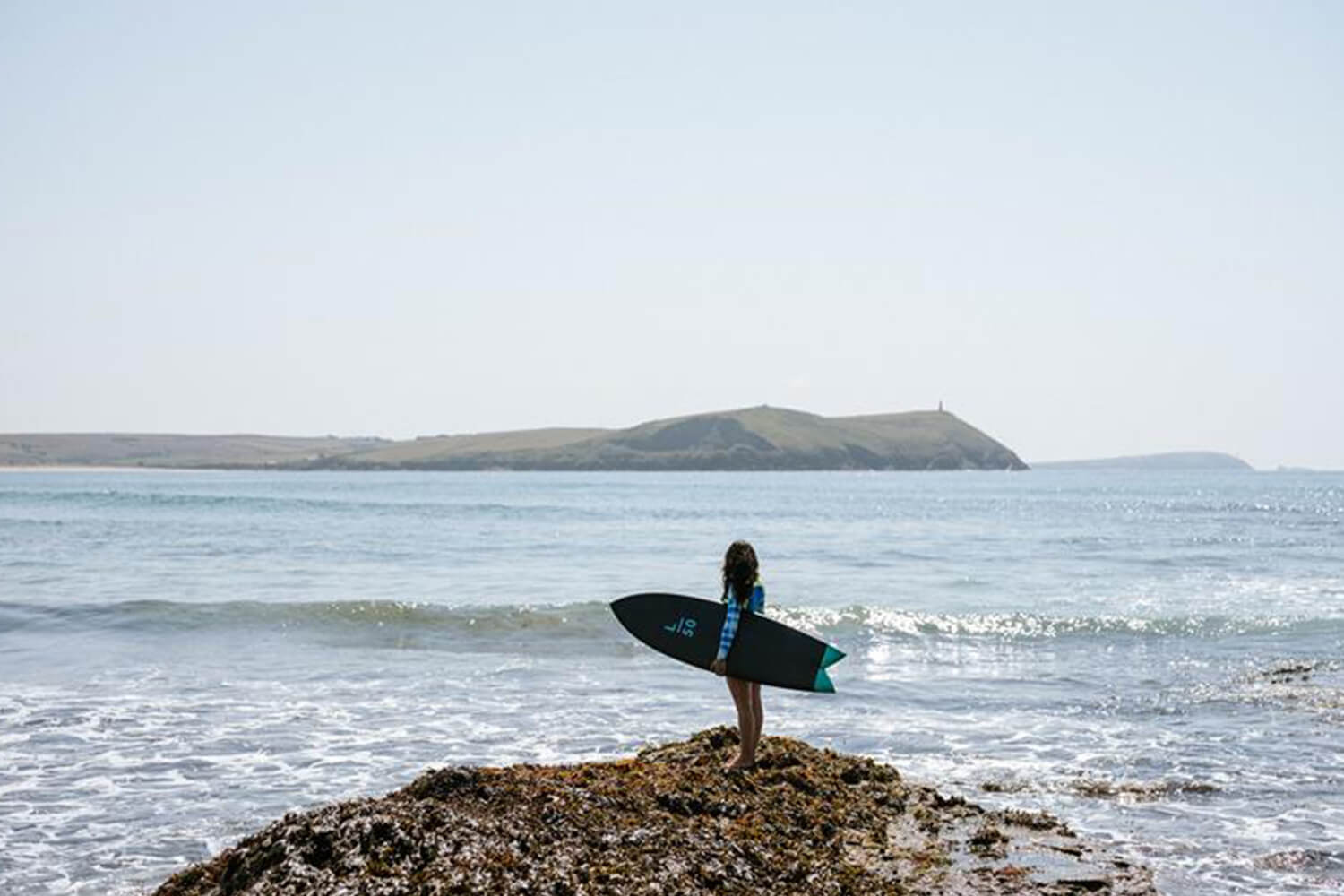 A female surfer stands on the shore looking out to sea with a surfboard under her arm.