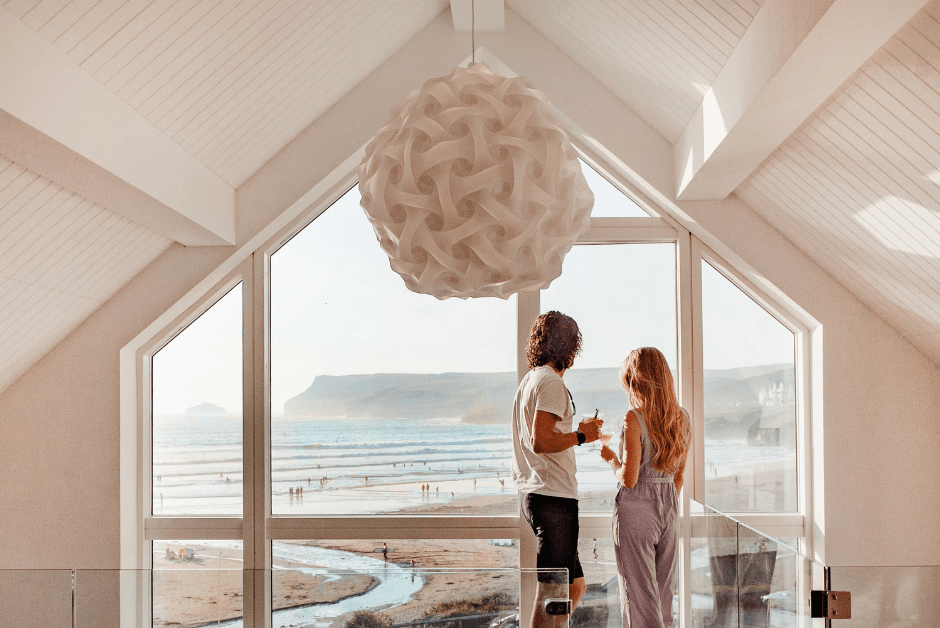 A man and woman look out over the beach through the large floor-to-ceiling window of Chyanna in Polzeath