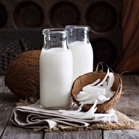 two glass bottles of coconut milk with a coconut shell filled with coconut flakes