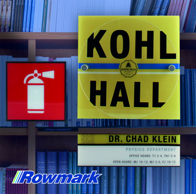 Rowmark Slickers engraved plastic signage including fire safety, hall identification, and office nameplate signs displayed on bookshelves