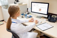 Back view portrait of blonde female doctor sitting at desk in office working with computer, typing and looking at blank white screen, copy space