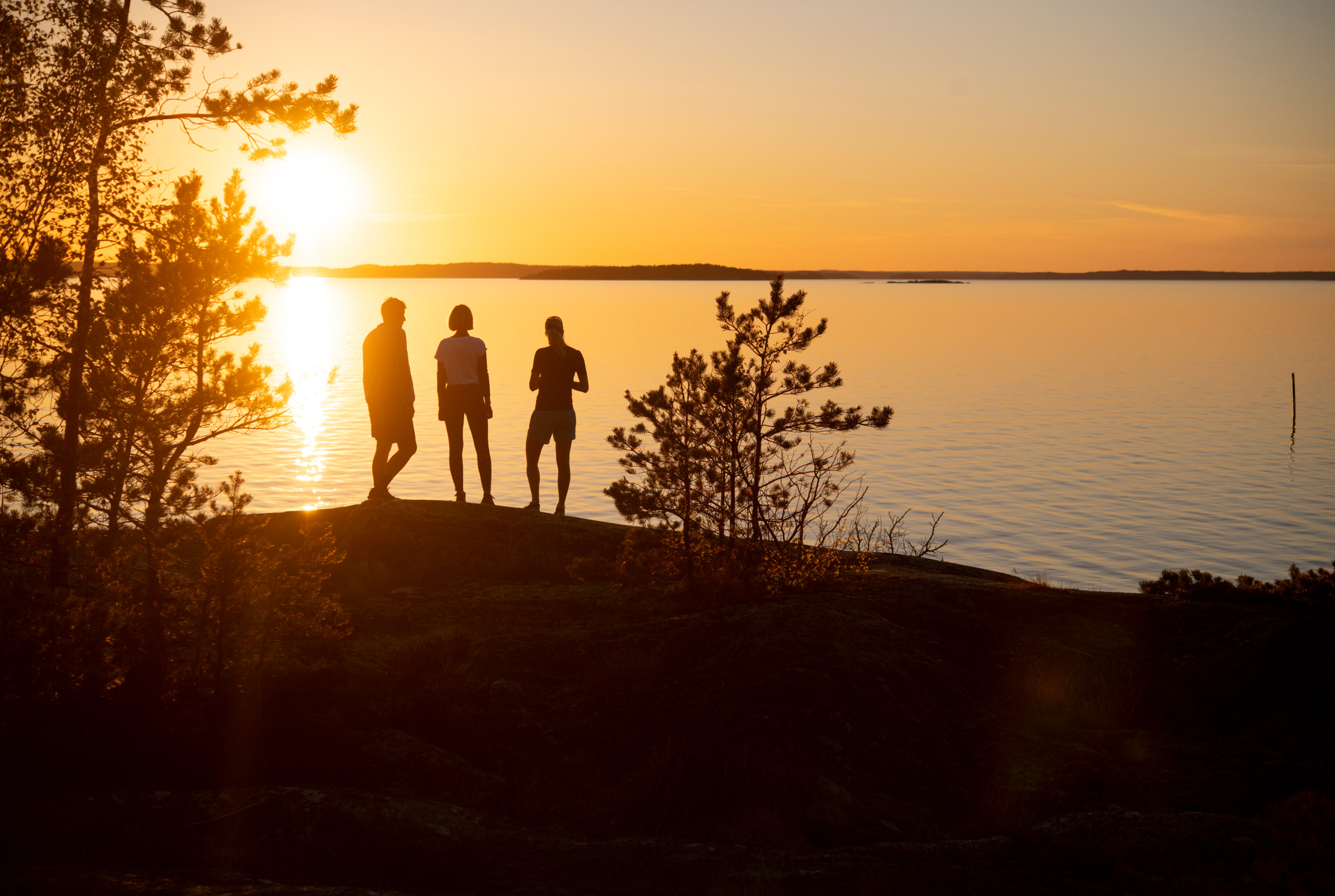 Three people are standing by the sea and looking at the sunset.