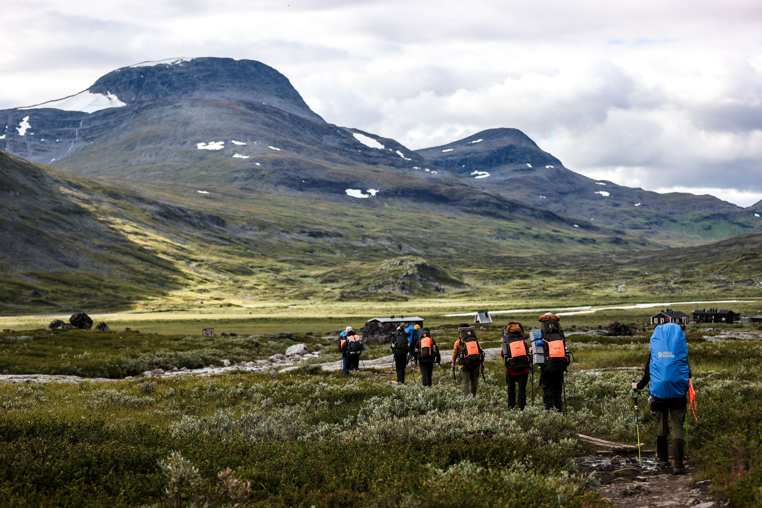A group of people hiking Kungsleden, with a majestic background of mountains, cloudy skies, fields, sunshine.