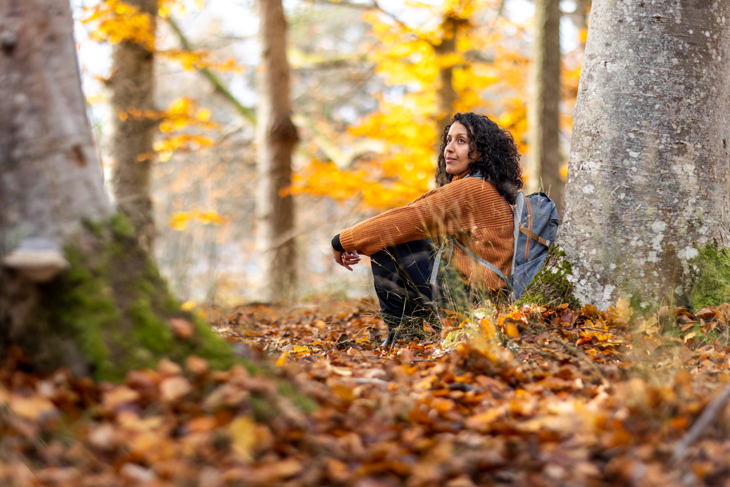 En kvinna sitter lutad mot ett träd i skogen och tar det lugnt. Det är höst och marken är täckt av löv och solen strilar genom trädens grenar.