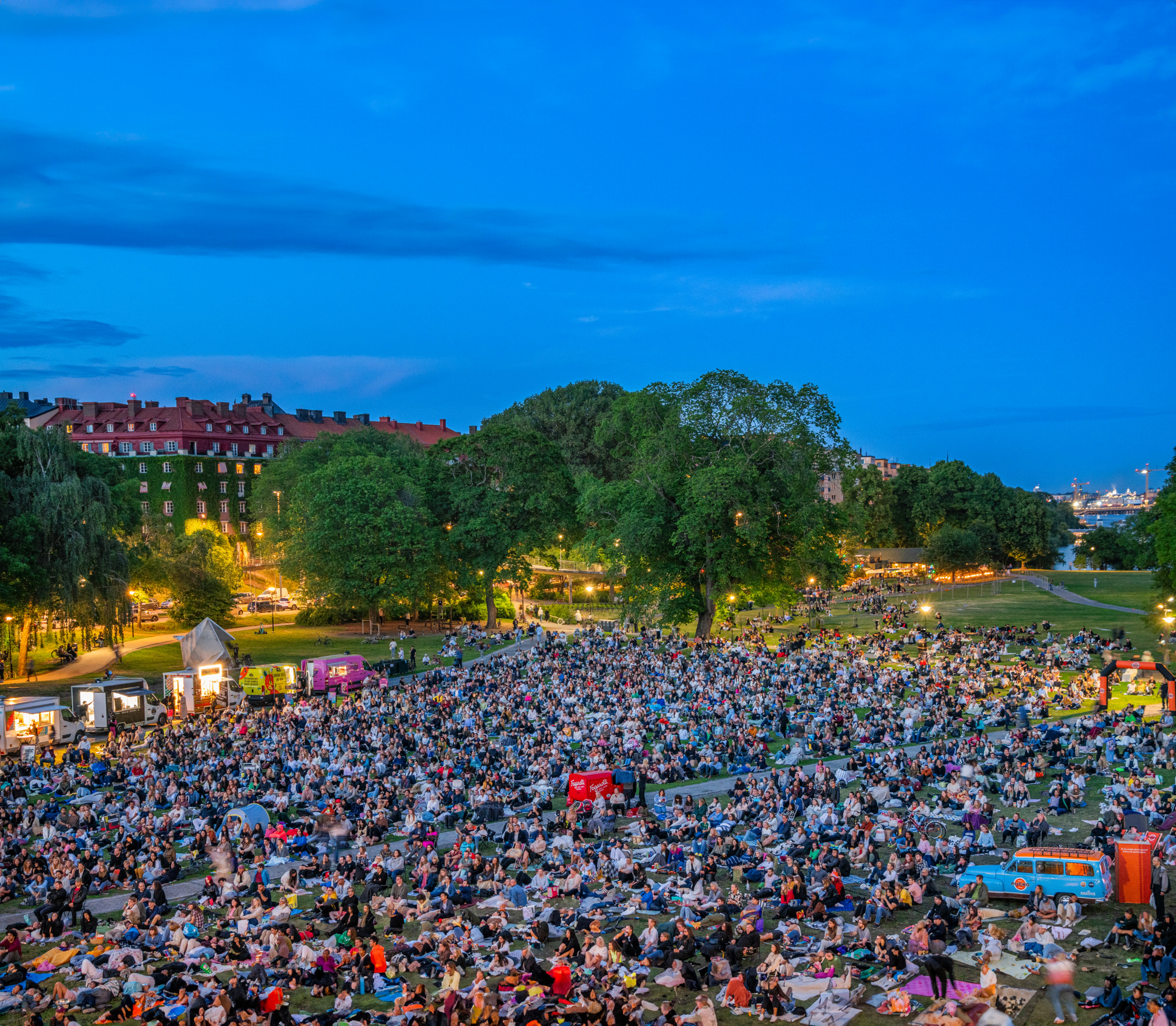 En gräsmatta full av människor en sommarkväll i Stockholm. Alla tittar på utomhusbio. Runt gräsmattan står det flera food-trucks.